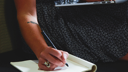Woman in dress sittingwith laptpo on lap but using a pen to write on a notepad next to her on the sofa